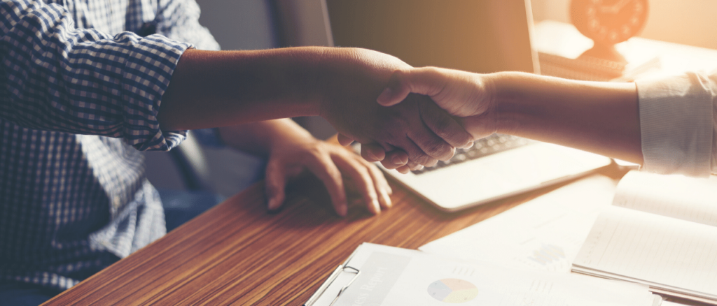 Two people shaking hands across a desk which has various papers and a laptop.